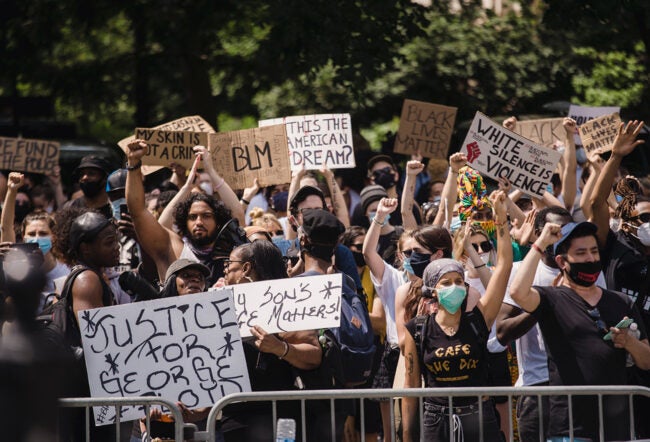 A large group of protesters hold signs advocating for racial justice and equality. Some signs read "BLM," "My skin is not a crime," "Justice for George Floyd," and other messages promoting the Black Lives Matter movement. Protesters raise their fists