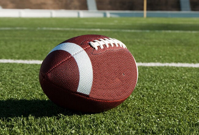 A football lies on a green field, representing the sport of American football. The scene includes white yard lines and a distant goalpost.